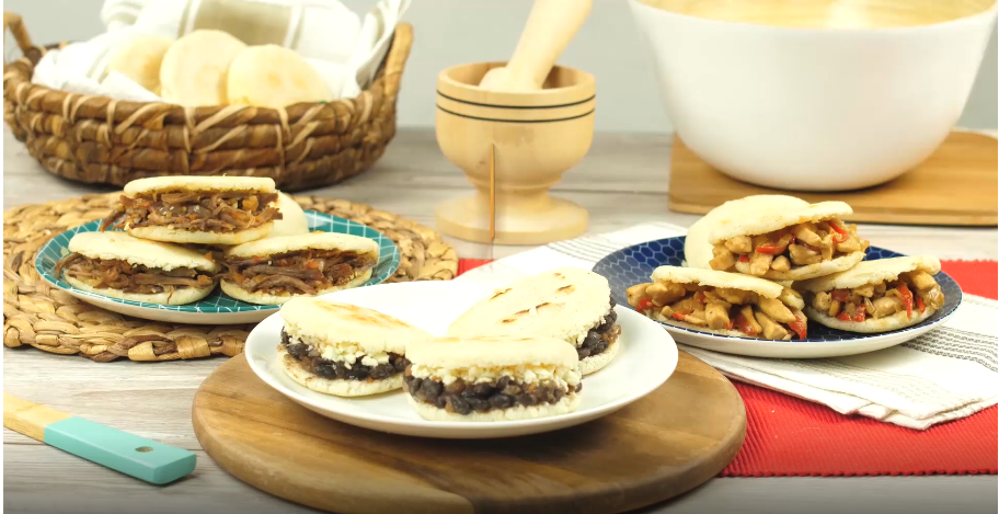 Arepas rellenas de pollo, frijoles negros y carne desmechada, sobre diferentes individuales. Al fondo se ven diferentes recipientes. Todo sobre una mesa de madera.