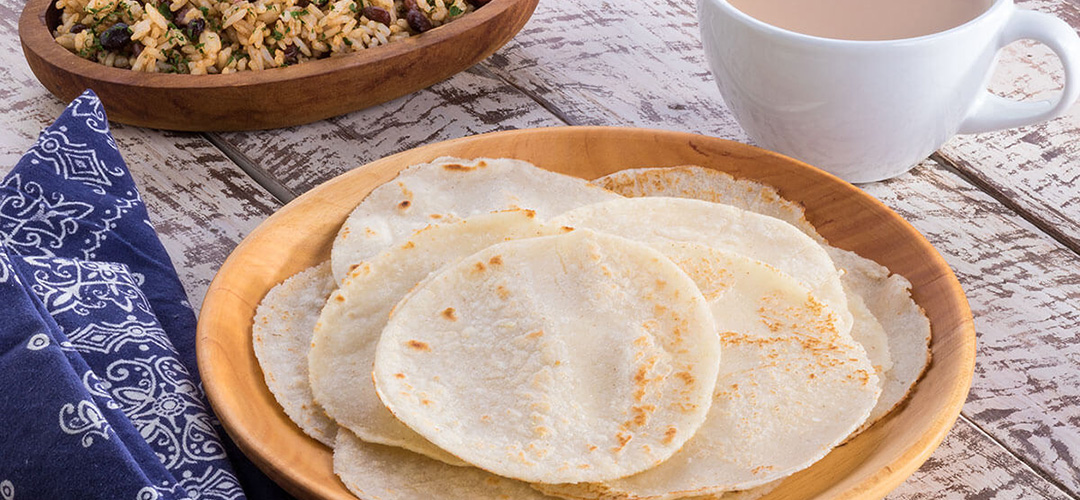 Tortillas servidas en un plato, al fondo se ve otro plato con arroz y al lado una taza de café y una servilleta de tela. Todo sobre una mesa de madera.