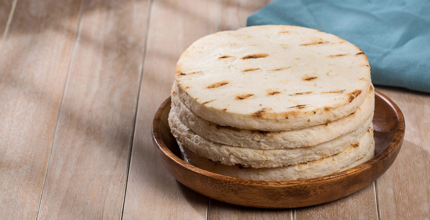 Torre de arepas de maíz asadas, servidas en un plato de madera. Las acompaña una servilleta de tela azul sobre una mesa de madera